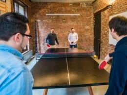 Vier erwachsene Männer spielen gemeinsam Tischtennis in einem modernen Büro mit großen Fenstern und Tageslicht.