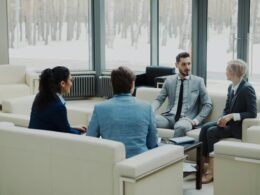 Vier Personen führen eine Besprechung in einem modernen, hell eingerichteten Büro mit großen Fenstern und Sitzgruppe.