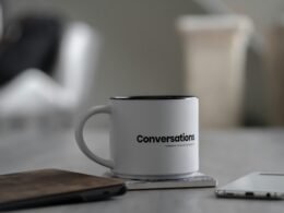 Weiße Tasse mit Chat-Symbolen neben einem Smartphone auf einem Schreibtisch im Büro- oder Homeoffice-Umfeld