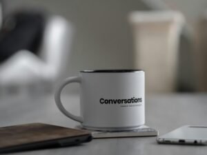 Weiße Tasse mit Chat-Symbolen neben einem Smartphone auf einem Schreibtisch im Büro- oder Homeoffice-Umfeld