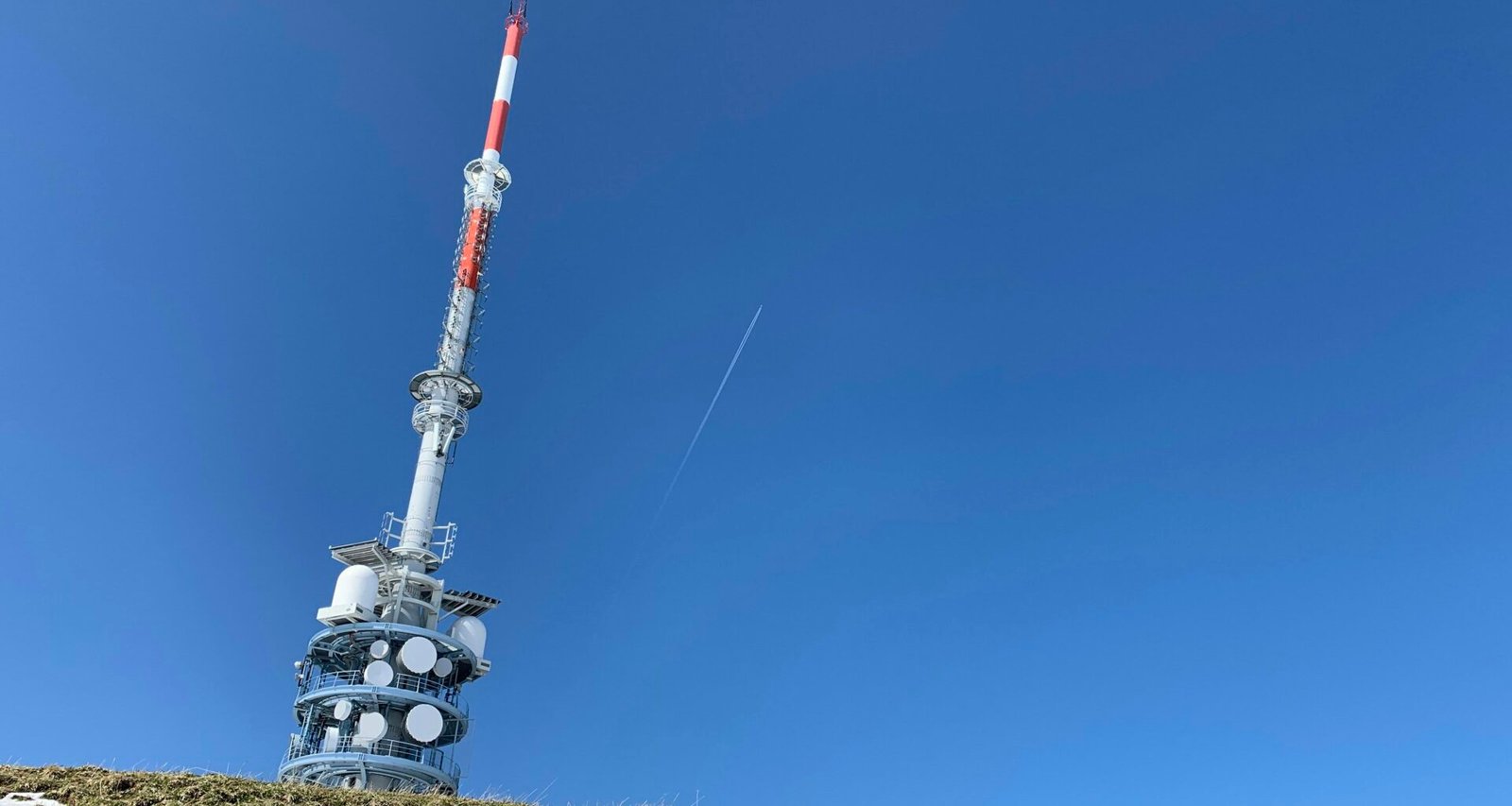 Weiß-roter Turm am Rand eines grünen Feldes, aufgenommen bei Tageslicht, neben einer kleinen Wiese, mit klarem Himmel