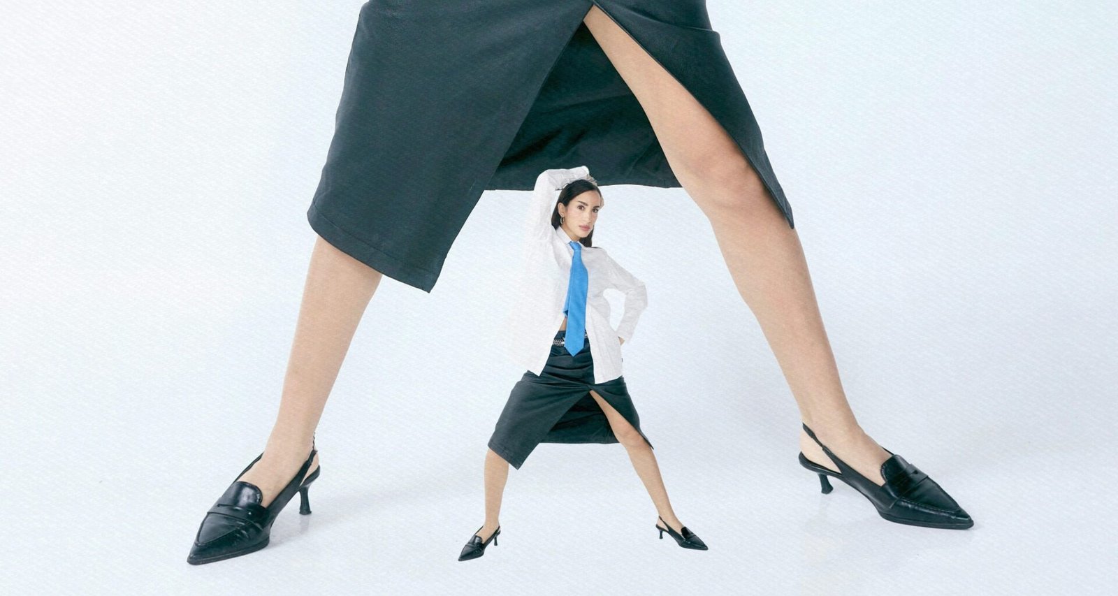 Eine winzige Frau steht zwischen zwei riesigen Beinen in langen Röcken und verdeutlicht deutliche Größenkontraste.