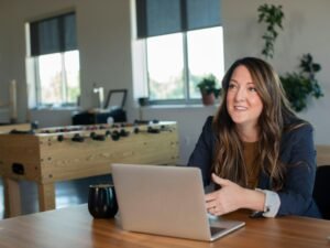 Eine Frau in brauner Strickjacke nutzt ein silbernes MacBook an einem Schreibtisch für ihre Online-Arbeit.