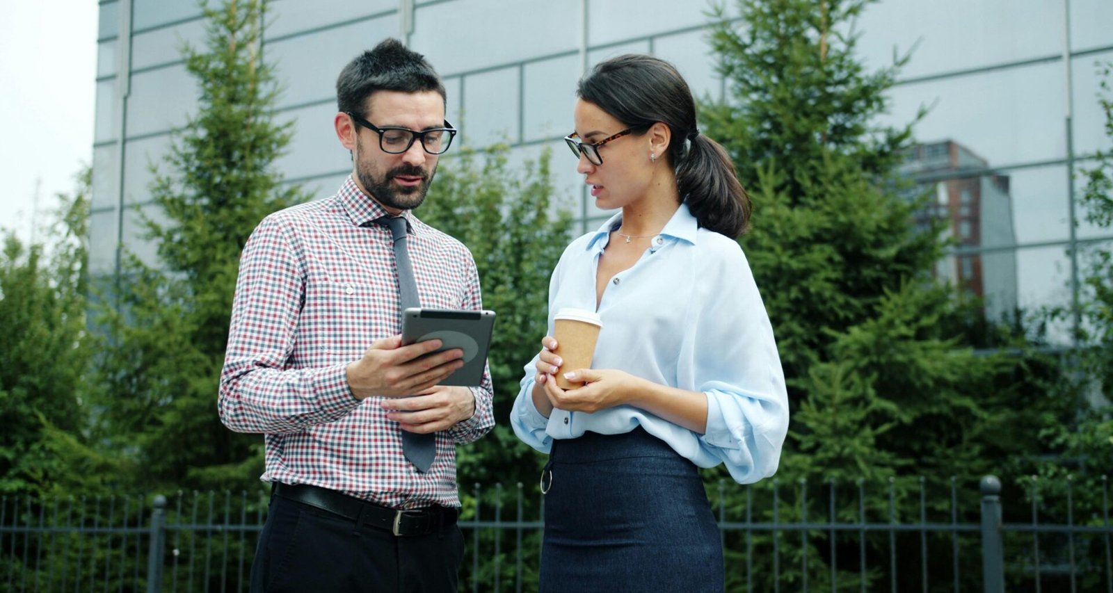 Zwei Berufstätige betrachten gemeinsam ein Tablet vor einem modernen Bürogebäude und diskutieren digitale Möglichkeiten.