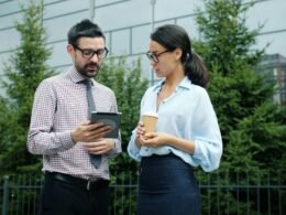Zwei Berufstätige betrachten gemeinsam ein Tablet vor einem modernen Bürogebäude und diskutieren digitale Möglichkeiten.