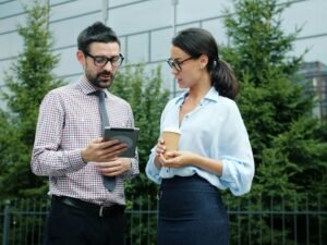 Zwei Berufstätige betrachten gemeinsam ein Tablet vor einem modernen Bürogebäude und diskutieren digitale Möglichkeiten.