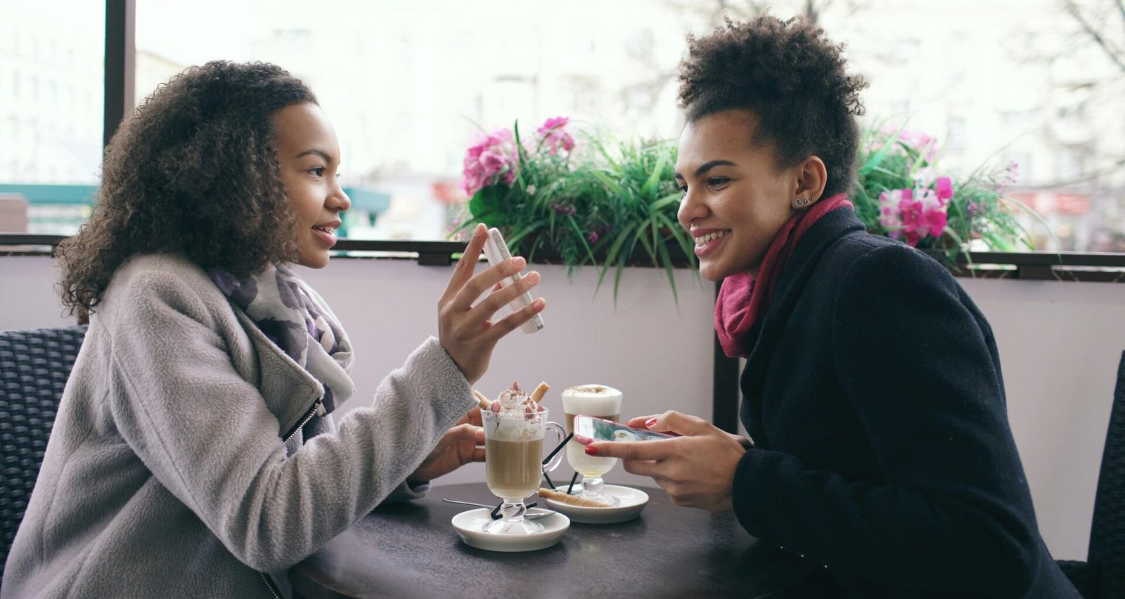Zwei Frauen sitzen an einem Tisch in einem Café und sprechen miteinander, im Hintergrund sind Getränke zu sehen.