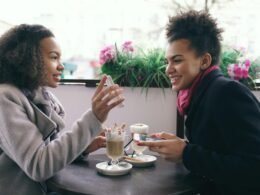 Zwei Frauen sitzen an einem Tisch in einem Café und sprechen miteinander, im Hintergrund sind Getränke zu sehen.