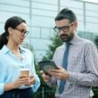 Zwei Geschäftspersonen betrachten und diskutieren gemeinsam Inhalte auf einem Tablet im Freien vor urbaner Kulisse.