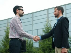 Zwei Männer im Anzug geben sich vor einem modernen Glasgebäude die Hand, im Freien, als Zeichen geschäftlicher Partnerschaft