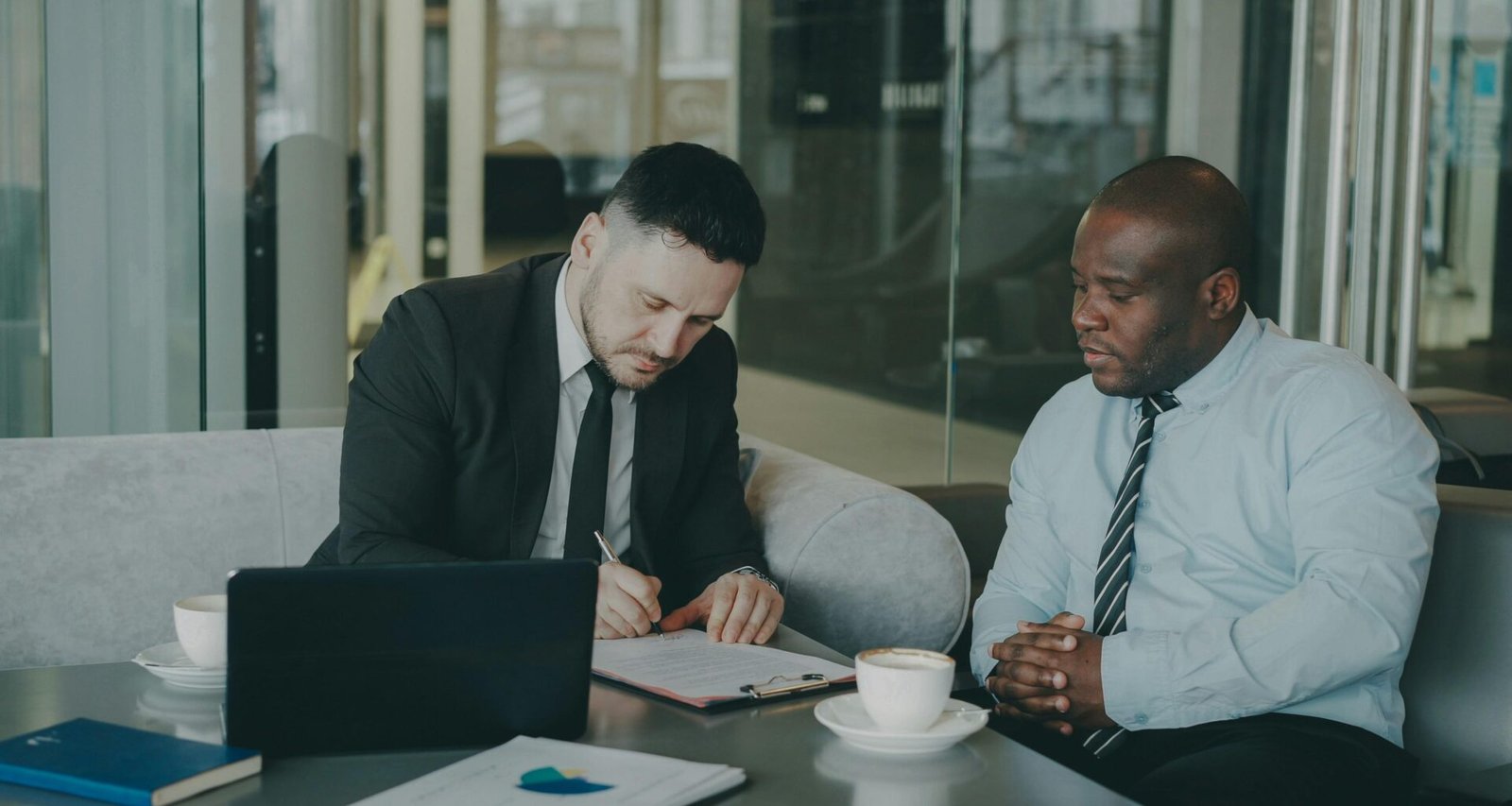 Zwei Männer im Business-Outfit unterschreiben gemeinsam einen Vertrag an einem Tisch in einem modern eingerichteten Büro