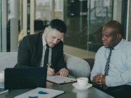 Zwei Männer im Business-Outfit unterschreiben gemeinsam einen Vertrag an einem Tisch in einem modern eingerichteten Büro