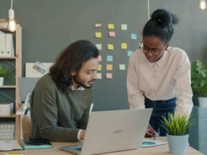 Zwei Kollegen sitzen an einem Schreibtisch und besprechen gemeinsam ein Projekt mit Laptop und Unterlagen im Büro.