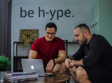 Zwei Männer sitzen an einem Couchtisch und schauen gemeinsam auf einen aufgeklappten Laptop im Büro.