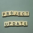 Zwei Scrabble-Steine mit den Worten project update liegen nebeneinander auf einem Holztisch und symbolisieren eine Projektaktualisierung.