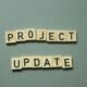 Zwei Scrabble-Steine mit den Worten project update liegen nebeneinander auf einem Holztisch und symbolisieren eine Projektaktualisierung.
